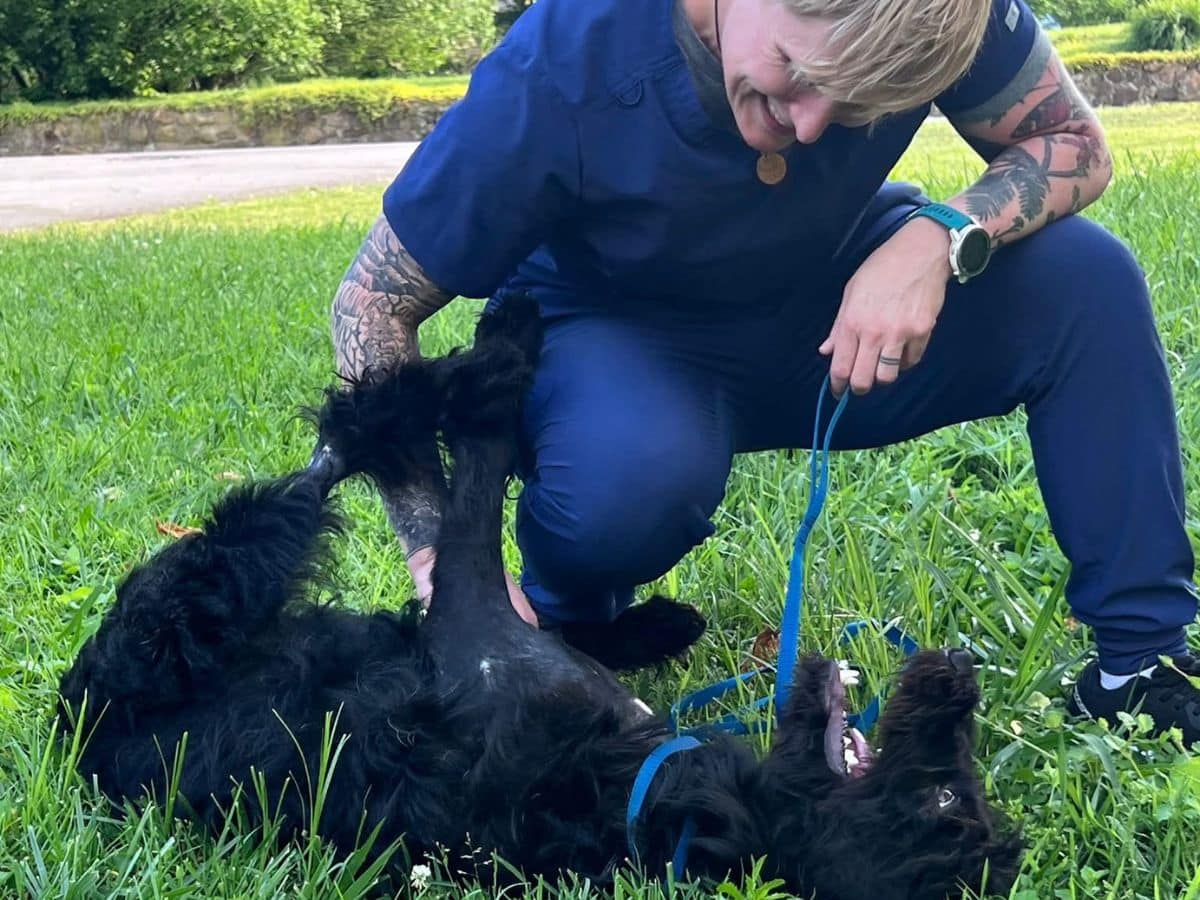 A woman kneels on grass beside a black dog